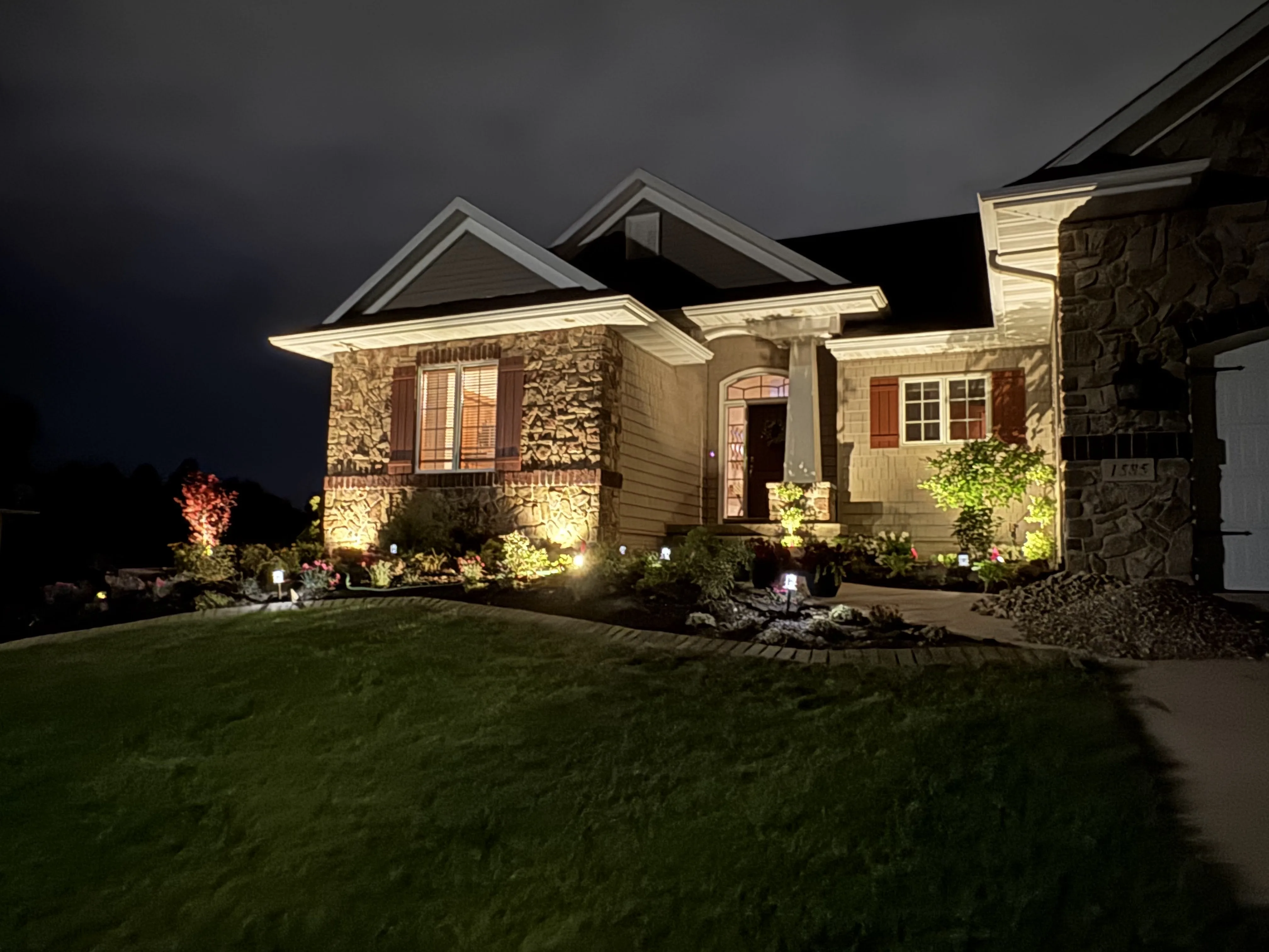 Front yard twilight landscape lighting with uplighting on stone home and plantings — Landforms Design Inc Eastern Iowa 2026