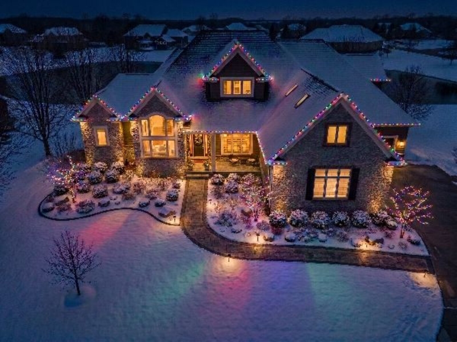 Robins Iowa front yard holiday lighting design preview by Darin Chamberlin — snowy scene with white roofline lights, multicolor uplighting sweeping across lawn and shrubs, festive door wreath